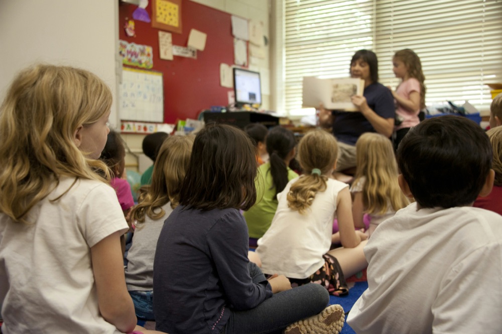 A teacher reading a picture book to a class of elementary school students sitting on the floor